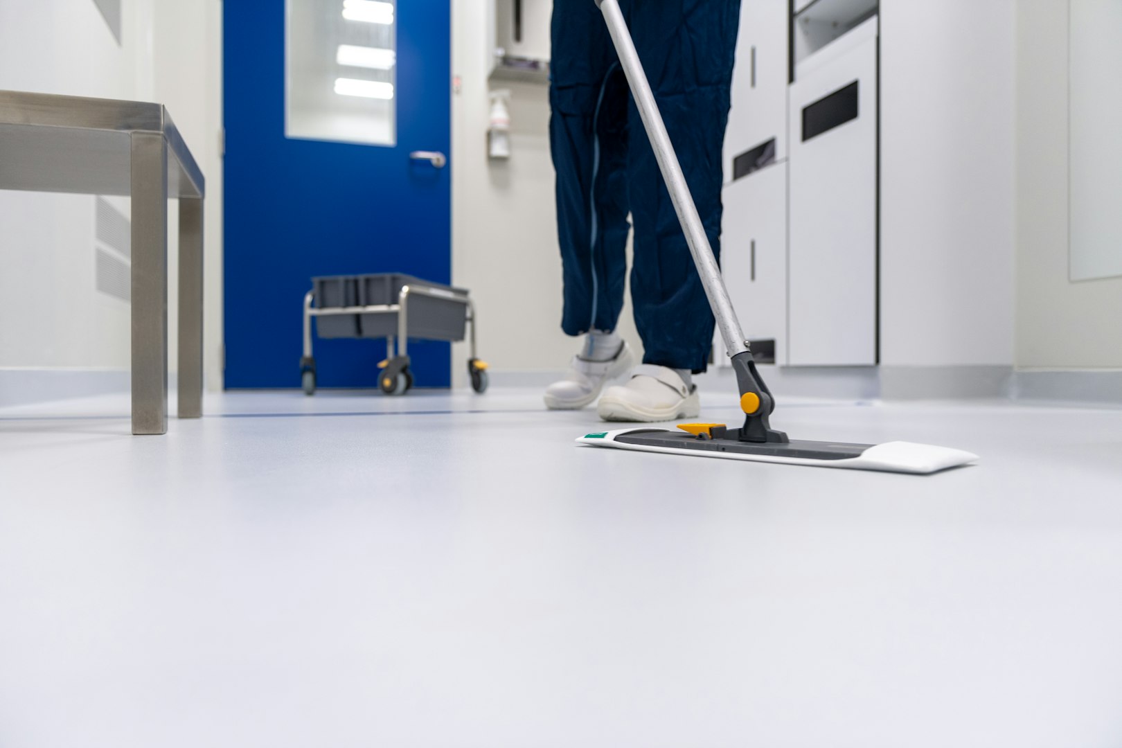 A person in work attire vacuuming a carpeted living room with a vacuum cleaner, featuring a large television in the background and a light-colored sofa.