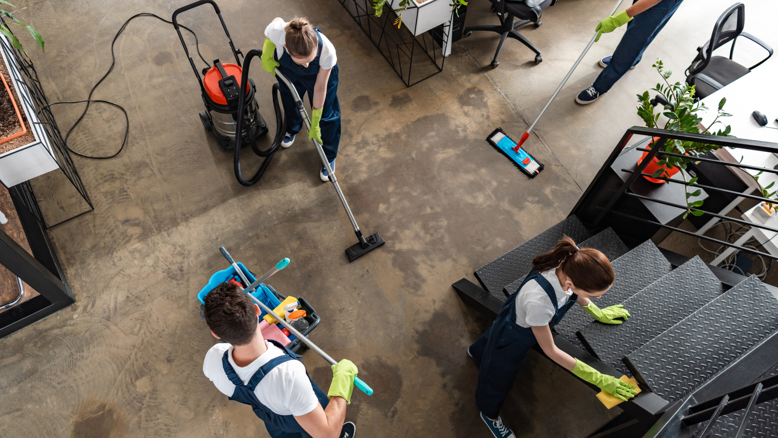 A group of four people in matching blue overalls and green gloves are cleaning an office space. Two individuals are using vacuum and mop equipment on the floor, while another person is cleaning stairs. Various plants and cleaning supplies are visible in the background.