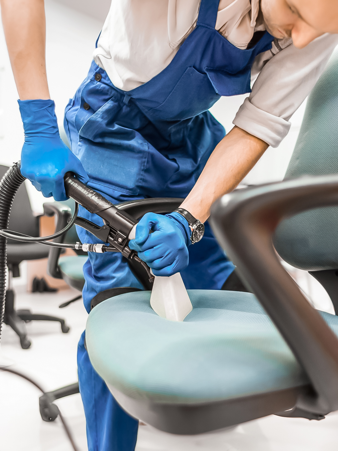 A person in blue overalls and gloves is using a cleaning machine to deep clean an office chair, focusing on the seat area.